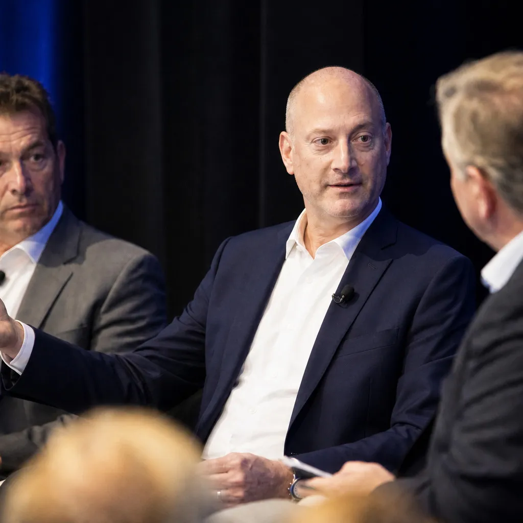 Peter DeCaprio speaking on a conference panel, gesturing as two fellow panelists listen